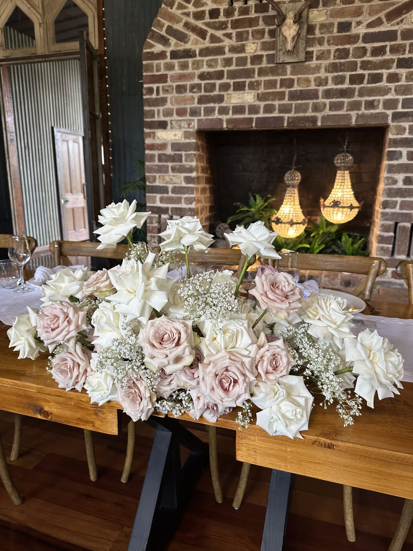 A long runner of white and dusty-pink roses down the reception table, framed by the barn's brick fireplace